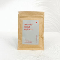 Kraft paper pouch of No-Nasty Bread Improver with its pale sage label reading softer loaves, simpler ingredients, standing on a white bench against a white wall