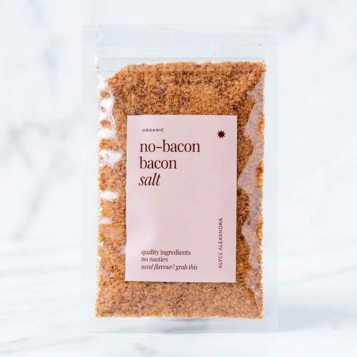 Alyce's No-Bacon Bacon Salt in a clear 100g packet with a soft pink label, the coarse smoky seasoning visible through the front