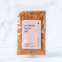 Alyce's No-Bacon Bacon Salt in a clear 100g packet with a soft pink label, the coarse smoky seasoning visible through the front