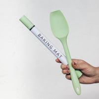 Hand holding a rolled Silicone Baking Mat in its tube beside a sage green spatula
