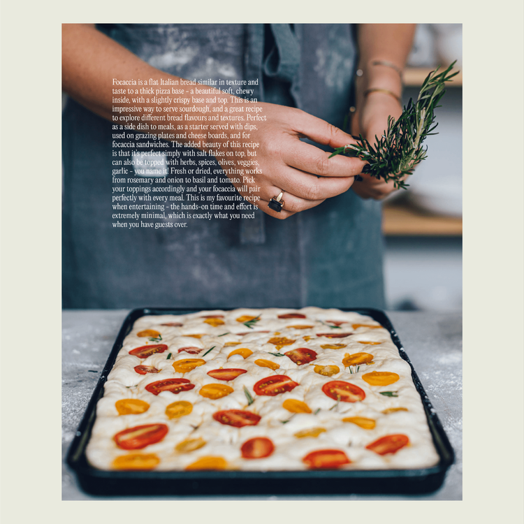 Hands scattering rosemary over a tray of sourdough focaccia topped with cherry tomatoes