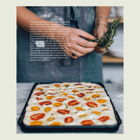 Hands scattering rosemary over a tray of sourdough focaccia topped with cherry tomatoes