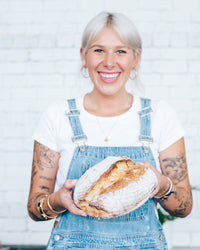 Ellen holding a large round sourdough loaf of bread in front of a white brick wall