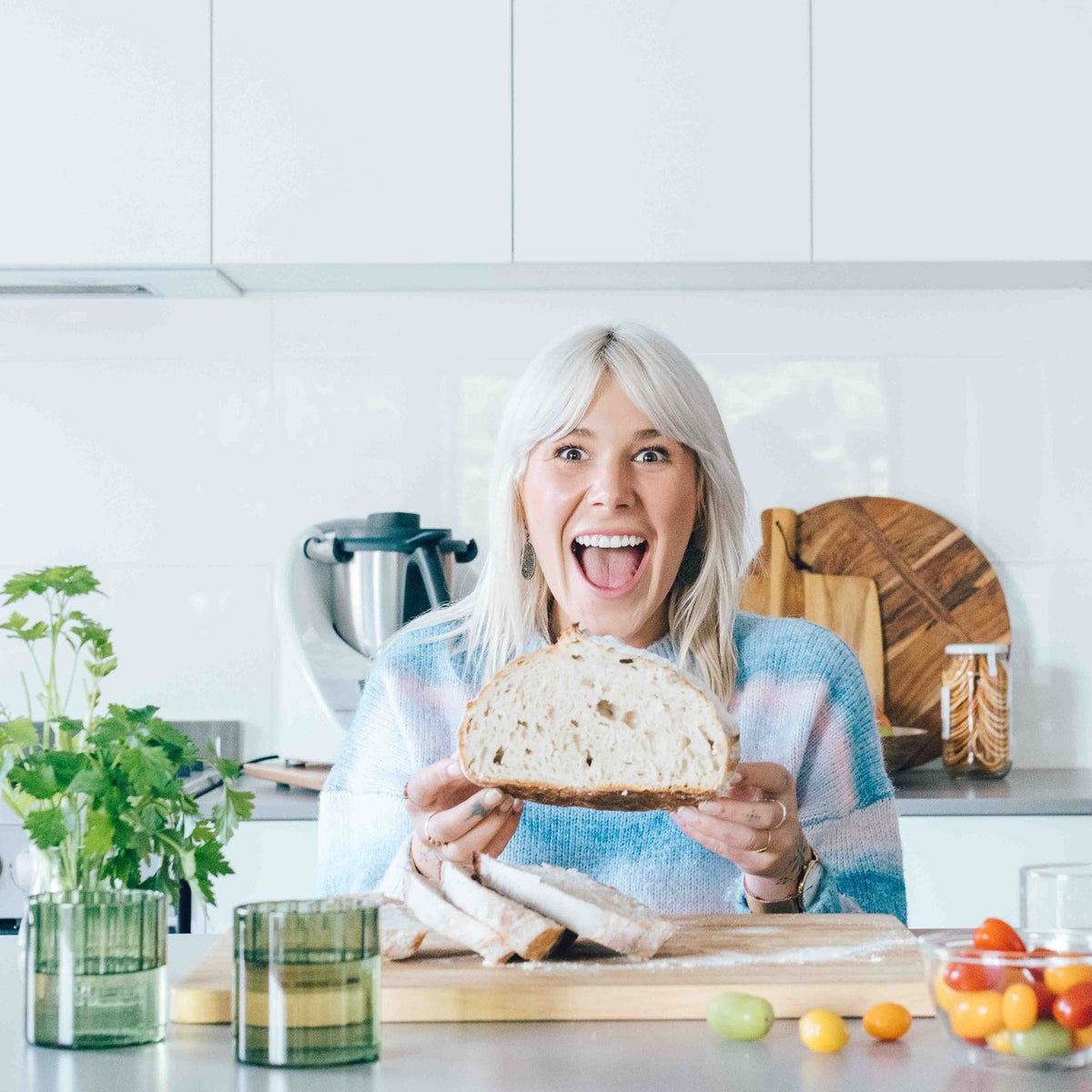 Alyce laughing in her kitchen holding a sourdough loaf of bread beside her Thermomix