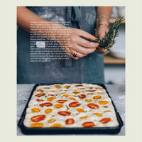 Hands scattering rosemary over a tray of sourdough focaccia topped with cherry tomatoes