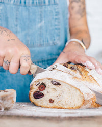 Hands slicing into a fruit-studded sourdough loaf of bread on a board