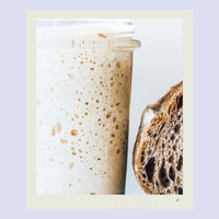 Jar of bubbling sourdough starter beside a dark sourdough loaf of bread