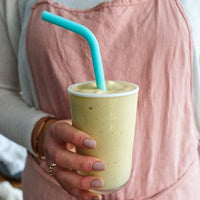 Hand holding a glass of green smoothie with a teal Wide Reusable Silicone Straw in it
