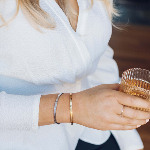 Woman in a white shirt holding a ribbed glass, wearing the gold 'You Are Amazing' Cuff alongside a fine chain bracelet