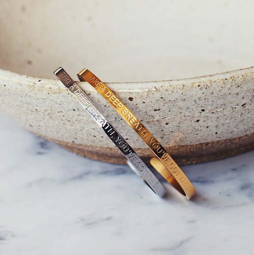 Three 'You've Got This' cuffs in rose gold, silver and gold leaning against a speckled ceramic bowl on marble, engraved take a deep breath, you've got this